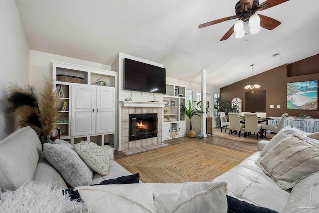 living room with ceiling fan with notable chandelier, a textured ceiling, lofted ceiling, wood-type flooring, and a tile fireplace