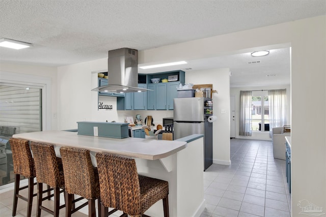 kitchen with stainless steel fridge, light tile patterned floors, a breakfast bar, island range hood, and blue cabinetry