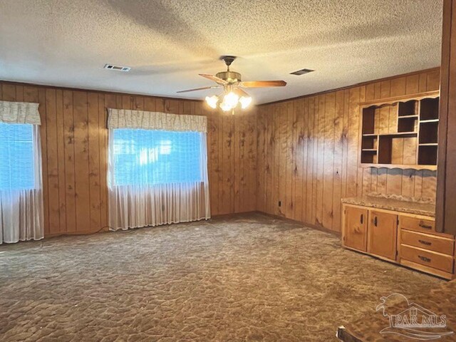 carpeted empty room featuring a wealth of natural light, wooden walls, a textured ceiling, and ceiling fan