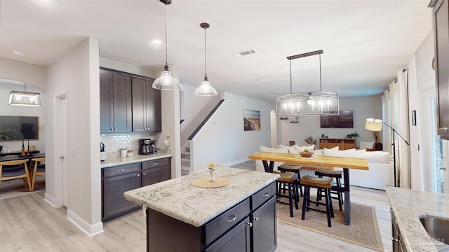 kitchen with dark brown cabinetry, a center island, decorative light fixtures, backsplash, and light stone counters