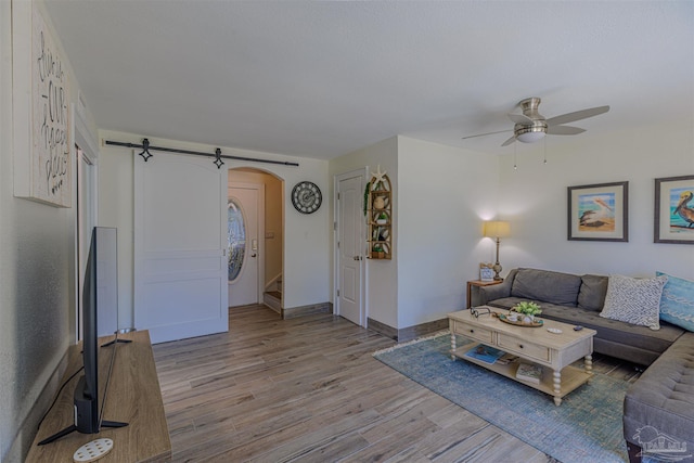 living room with arched walkways, light wood finished floors, a barn door, ceiling fan, and baseboards