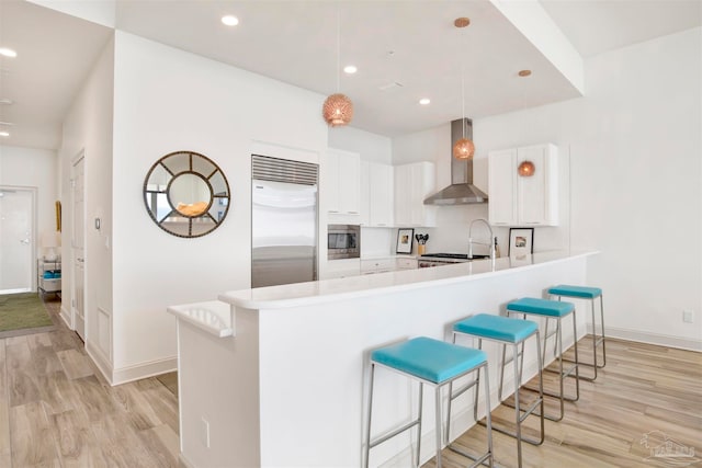 kitchen featuring white cabinetry, kitchen peninsula, built in appliances, decorative light fixtures, and wall chimney range hood