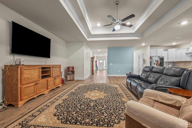 living room with baseboards, a raised ceiling, a ceiling fan, light wood-style flooring, and recessed lighting