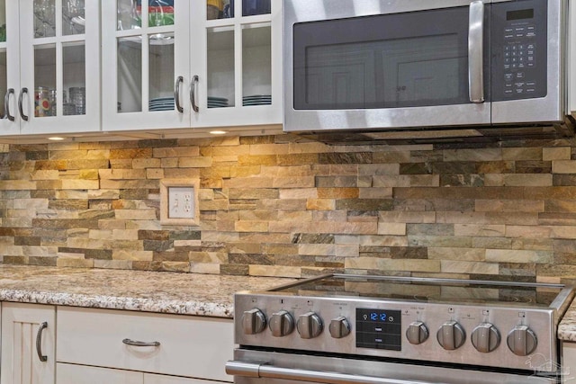 interior space with white cabinets, decorative backsplash, glass insert cabinets, light stone counters, and stainless steel appliances