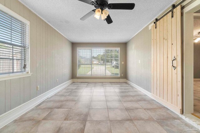 tiled empty room with wood walls, a textured ceiling, ceiling fan, and a barn door