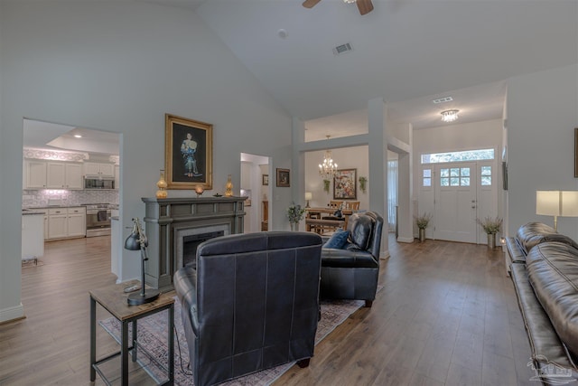 living room featuring a fireplace, high vaulted ceiling, ceiling fan with notable chandelier, wood finished floors, and visible vents