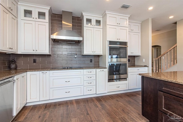 kitchen with decorative backsplash, glass insert cabinets, wall chimney exhaust hood, appliances with stainless steel finishes, and dark wood-type flooring