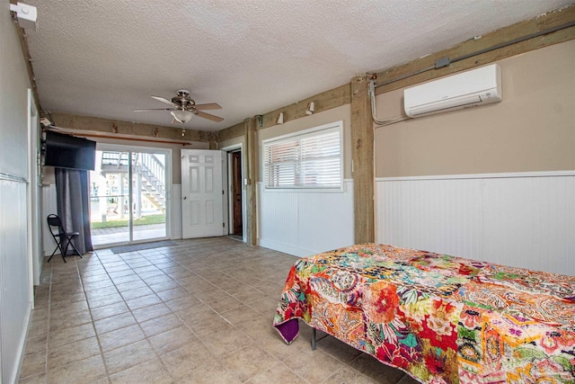 bedroom featuring a wall mounted air conditioner, a textured ceiling, wainscoting, ceiling fan, and access to exterior