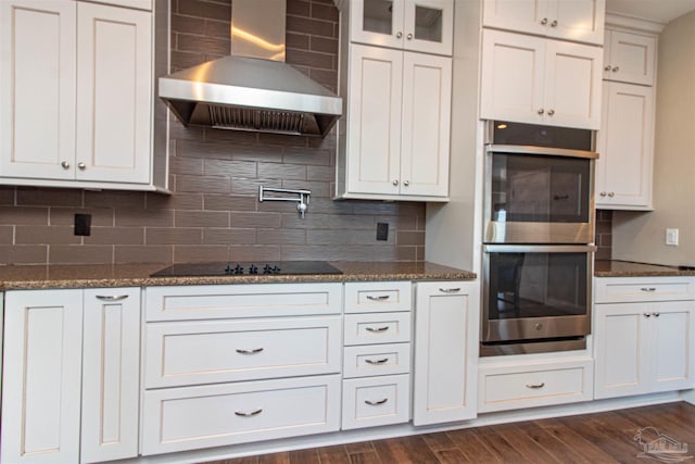 kitchen featuring dark stone countertops, double oven, dark hardwood / wood-style floors, wall chimney exhaust hood, and decorative backsplash