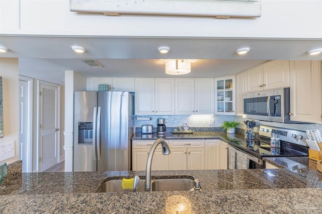 kitchen with dark stone counters, a sink, glass insert cabinets, appliances with stainless steel finishes, and backsplash