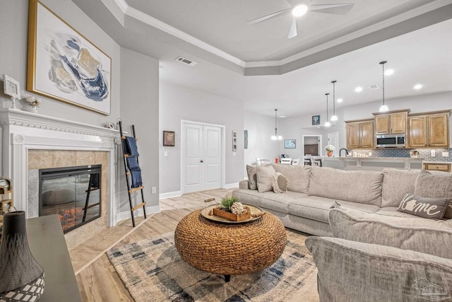 living room with crown molding, ceiling fan, a fireplace, a raised ceiling, and light wood-type flooring