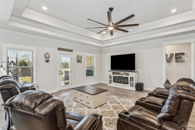 living area featuring light wood-style flooring, a fireplace, a ceiling fan, a raised ceiling, and crown molding