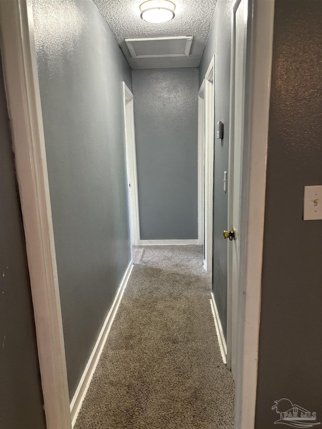 hallway featuring baseboards, attic access, carpet flooring, a textured wall, and a textured ceiling