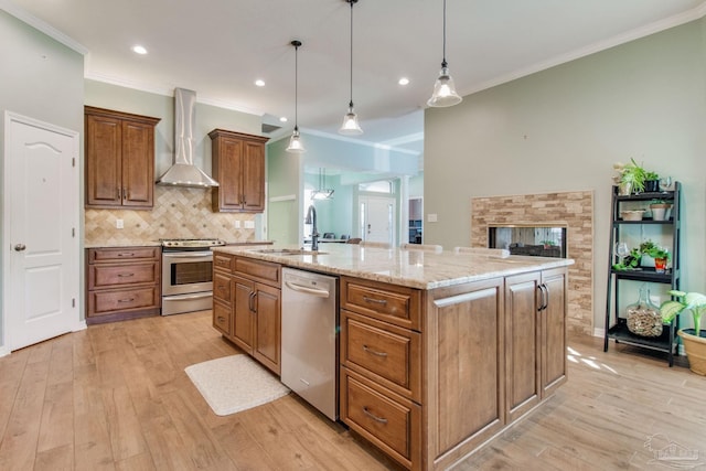 kitchen with a center island with sink, stainless steel appliances, sink, wall chimney range hood, and decorative light fixtures