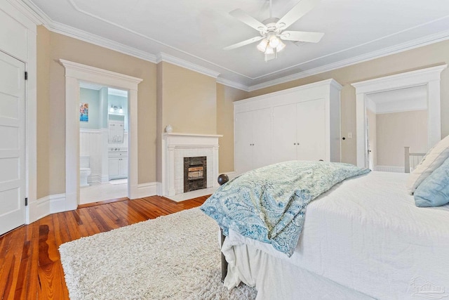 bedroom featuring hardwood / wood-style flooring, connected bathroom, crown molding, a brick fireplace, and ceiling fan