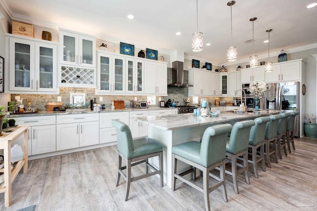 kitchen with light hardwood / wood-style flooring, white cabinetry, wall chimney exhaust hood, an island with sink, and ornamental molding