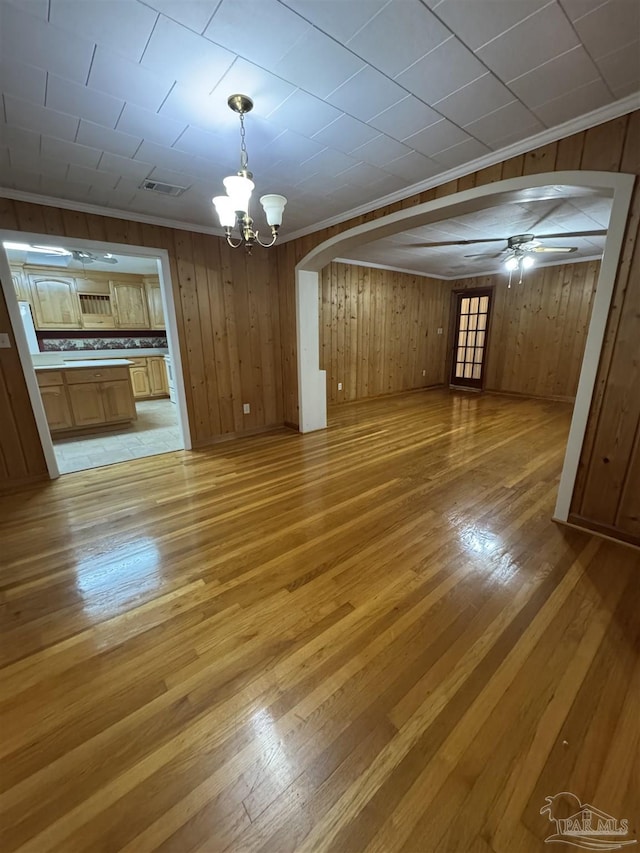 unfurnished living room featuring ceiling fan with notable chandelier, ornamental molding, wood walls, and light wood-type flooring