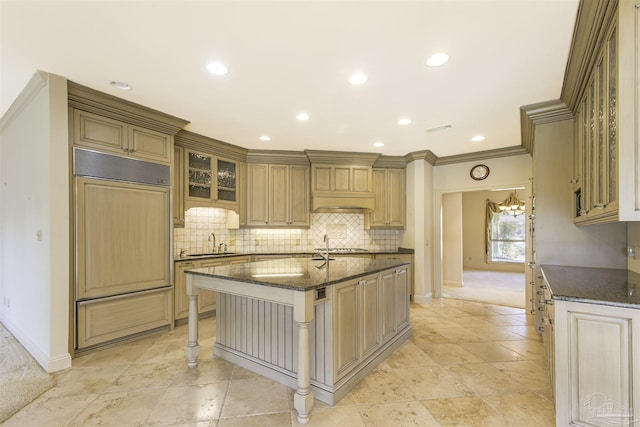 kitchen with sink, crown molding, dark stone countertops, paneled built in fridge, and an island with sink