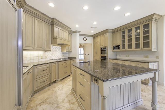 kitchen with appliances with stainless steel finishes, tasteful backsplash, sink, dark stone counters, and a kitchen island with sink