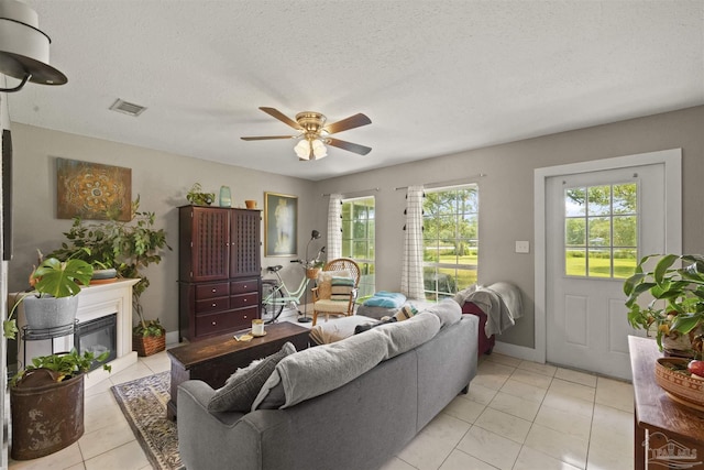 living room featuring visible vents, light tile patterned floors, a glass covered fireplace, a textured ceiling, and a ceiling fan