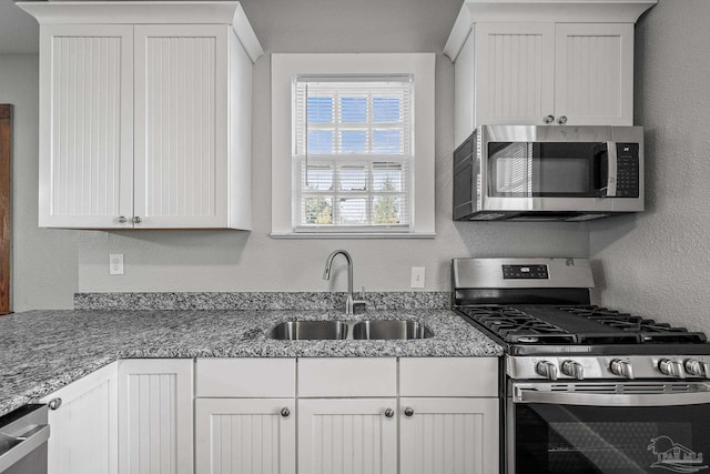 kitchen with a textured wall, stone counters, stainless steel appliances, a sink, and white cabinets