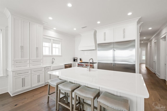 kitchen with white cabinets, an island with sink, premium range hood, dark hardwood / wood-style flooring, and premium appliances