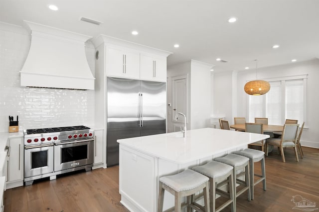 kitchen with custom exhaust hood, high quality appliances, white cabinets, and an island with sink