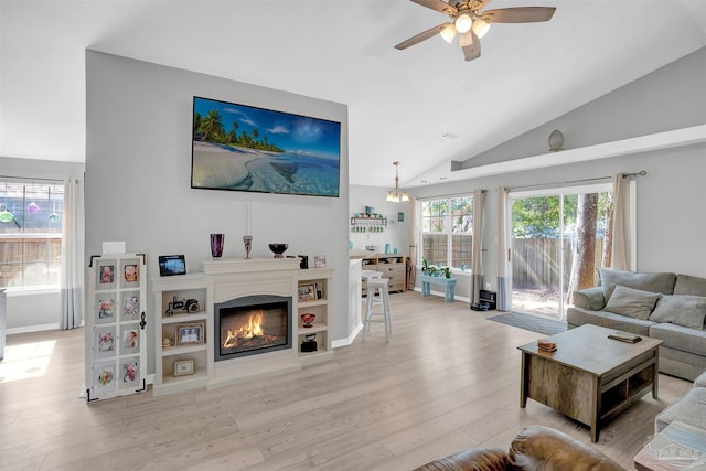 living area featuring ceiling fan with notable chandelier, baseboards, vaulted ceiling, light wood-style floors, and a glass covered fireplace