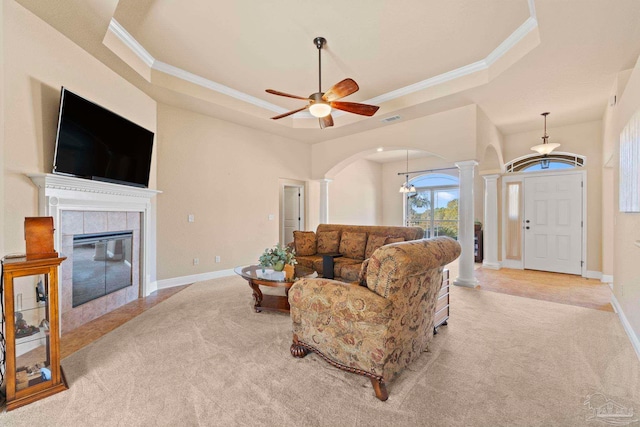 living room with a tile fireplace, a raised ceiling, ceiling fan, ornate columns, and ornamental molding