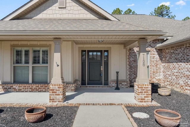 view of exterior entry with a porch, brick siding, and roof with shingles