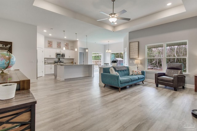 living room featuring baseboards, a tray ceiling, recessed lighting, ceiling fan with notable chandelier, and light wood-type flooring