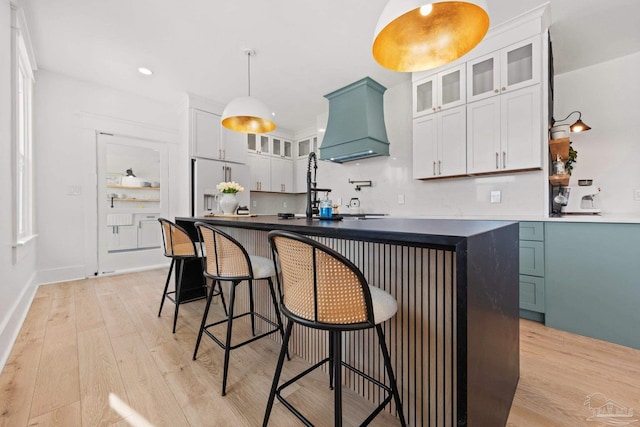 kitchen featuring light wood finished floors, dark countertops, custom range hood, high end refrigerator, and a kitchen island with sink