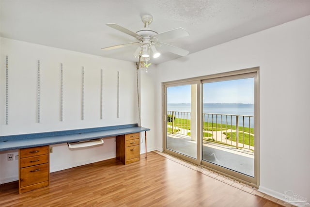 unfurnished office featuring a water view, light hardwood / wood-style flooring, ceiling fan, built in desk, and a textured ceiling