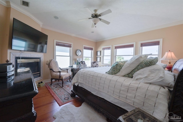 bedroom with a tile fireplace, wood finished floors, visible vents, a ceiling fan, and ornamental molding