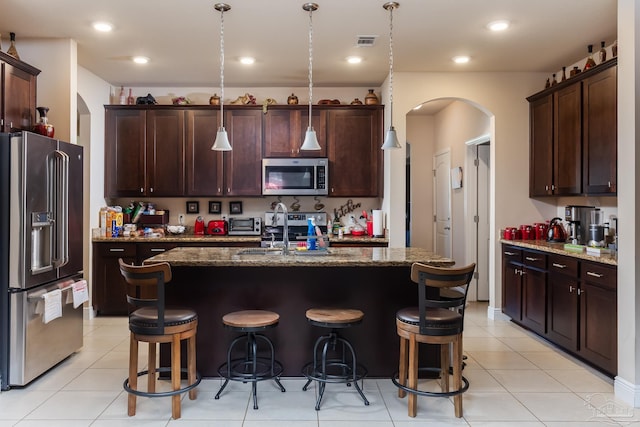 kitchen featuring arched walkways, appliances with stainless steel finishes, a kitchen island with sink, and decorative light fixtures