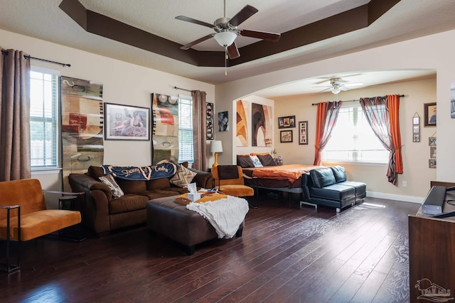 living room with ceiling fan, arched walkways, dark wood finished floors, baseboards, and a raised ceiling