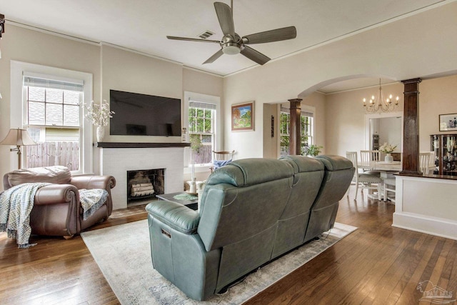 living area with decorative columns, hardwood / wood-style flooring, arched walkways, a brick fireplace, and ceiling fan with notable chandelier