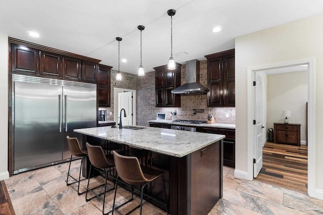kitchen with wall chimney exhaust hood, stainless steel appliances, a sink, stone tile floors, and dark brown cabinetry