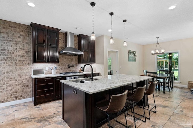 kitchen featuring wall chimney exhaust hood, a sink, a center island with sink, baseboards, and light stone counters