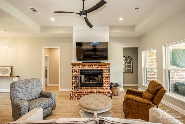 living room with a tray ceiling, a brick fireplace, light wood-type flooring, and ceiling fan