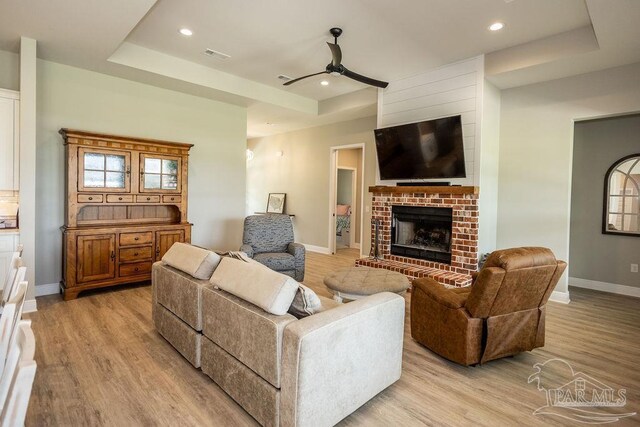 living room with ceiling fan, light hardwood / wood-style floors, a brick fireplace, and a tray ceiling