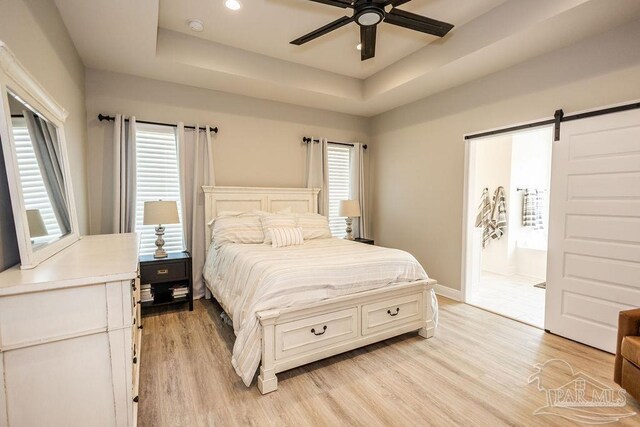 bedroom with light hardwood / wood-style flooring, connected bathroom, a barn door, ceiling fan, and a tray ceiling