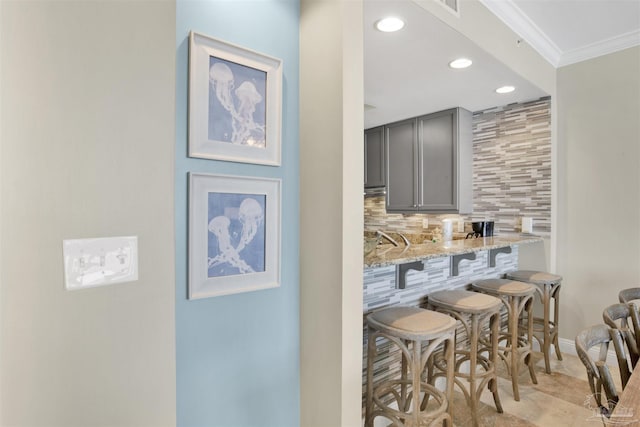 kitchen featuring gray cabinets, a kitchen bar, decorative backsplash, ornamental molding, and light stone counters