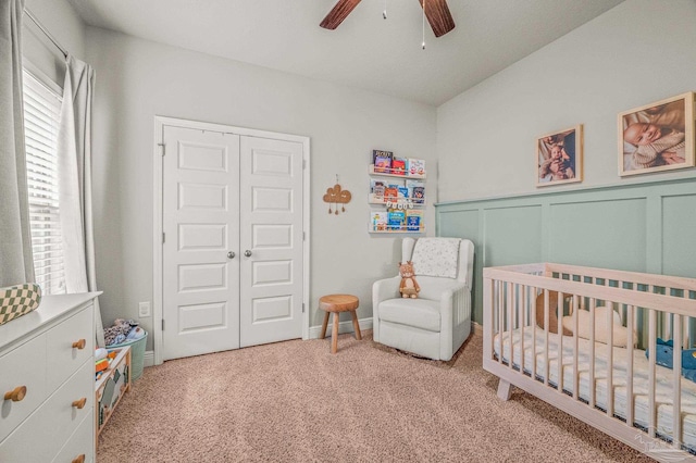 carpeted bedroom featuring a crib, ceiling fan, a closet, and a wainscoted wall