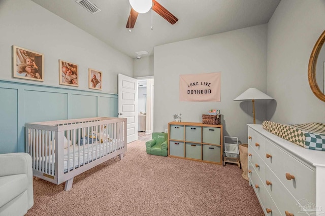 carpeted bedroom with a crib, a ceiling fan, a decorative wall, and wainscoting