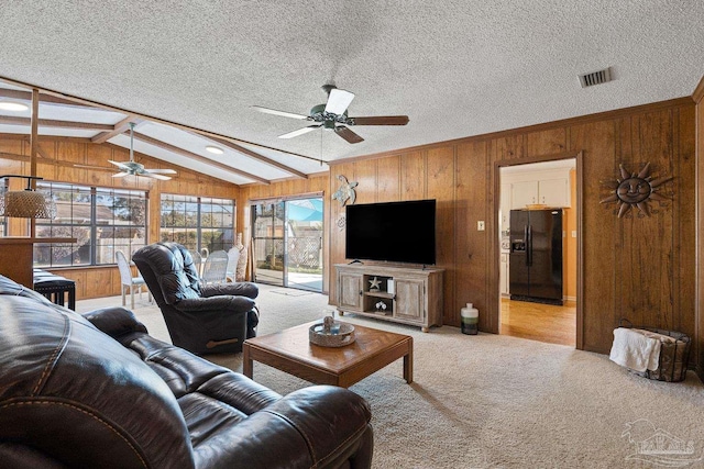 living room with wooden walls, vaulted ceiling with beams, light colored carpet, ceiling fan, and a textured ceiling