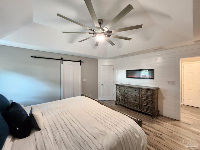 bedroom with light wood-type flooring, a raised ceiling, ceiling fan, a barn door, and wood walls