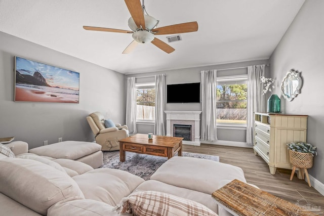 living area with visible vents, light wood-style flooring, a glass covered fireplace, ceiling fan, and baseboards