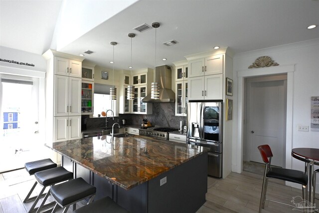 kitchen with tasteful backsplash, stainless steel appliances, a kitchen island with sink, wall chimney range hood, and hanging light fixtures