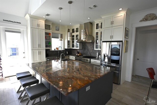 kitchen featuring wall chimney exhaust hood, hanging light fixtures, tasteful backsplash, a center island with sink, and appliances with stainless steel finishes